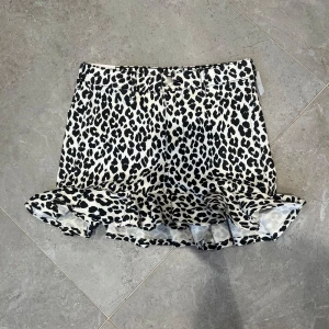 Leopardmönstrad kortkjol med volang - Snygg kortkjol med svartvitt leopardmönster och volangkant nertill. Kjolen har klassisk jeansmodell med fickor bak och bälteshällor. Perfekt statement-plagg för dig som vill sticka ut.