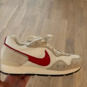 Nike sneakers med röd swoosh - Snygga Nike sneakers i beige och vitt med klassisk röd swoosh på sidorna. Ovandelen är i mesh och mocka, med vita snören och svart sula. Retroinspirerad design som passar perfekt till streetwear. Sköna och lätta på foten.