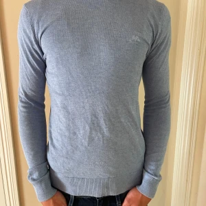 Ljusblå stickad tröja från Lindbergh - Snygg ljusblå stickad tröja från Lindbergh med diskret logga på bröstet. Tröjan har rund halsringning, ribbade muddar och är långärmad. Perfekt för dig som gillar en clean och stilren look.