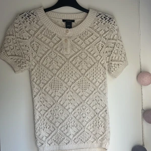 Beige stickad topp från Lindex - Säljer en beige stickad topp från Lindex med korta ärmar och snyggt hålmönster. Tröjan har rund halsringning, ribbade kanter och två dekorativa knappar framtill. Perfekt för dig som gillar en chill och trendig stil.