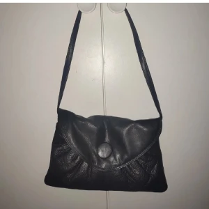 Depesche Svart axelväska i skinn - Snygg svart axelväska i mjukt skinn med lock och stor knapp framtill. Väskan har en enkel, klassisk design med lätt rynkade detaljer på framsidan och en lång axelrem. Perfekt för att bära det viktigaste när du är på språng.