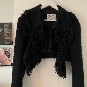 Svart croppad jacka med fransar Zara - Svart croppad jacka från Zara Studio med långa ärmar och coola fransdetaljer framtill, på axlarna och längs ryggen. Jackan har en grovstickad känsla och bred krage, perfekt för dig som vill sticka ut med en edgy look.
