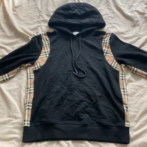 Burberry huvtröja - Svart huvtröja från Burberry med klassiska beige-rutiga paneler längs sidor och ärmar. Tröjan har huva med snörning och en stor magficka. Perfekt för dig som vill ha en streetig och lyxig vibe i garderoben.