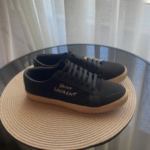 Svarta Saint Laurent sneakers - Svarta sneakers från Saint Laurent med beige sula och handskriven logga på sidan. Skorna har snörning, rund tå och är tillverkade i en mix av skinn och textil. Perfekta för dig som gillar stilrena och exklusiva sneakers med en unik touch.