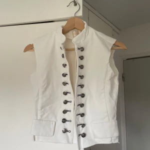 Vit väst med dekorativa knappar - Snygg vit väst från med ståkrage och dubbla rader av dekorativa metallknappar framtill. Västen har fickdetaljer och en stilren, lite militärinspirerad look. Perfekt att styla över en skjorta eller t-shirt för en cool outfit.