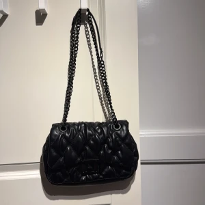 Svart quiltad axelväska med kedja - Säljer en svart quiltad axelväska i skinnimitation med chunky kedjerem. Väskan har lock med snyggt spänne framtill och coolt mönstrad yta. Perfekt storlek för mobilen, plånboken och lite till. Kedjan ger en edgy vibe till hela looken.