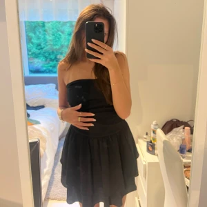 Svart axelbandslös kortklänning - Supersnygg svart axelbandslös klänning med volangig kjol och figurnära överdel. Klänningen har en kort längd och en luftig, flowy känsla som passar perfekt för en trendig look. Enkel att styla med smycken och klackar för en extra touch.