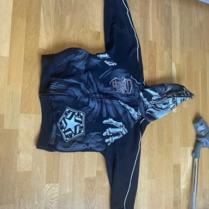 Svart hel zipupjacka Tribal Gear - Svart hoodiejacka från Tribal Gear med grått och vitt tryck på framsidan, inklusive skelettmotiv och logga. Jackan har hel dragkedja, huva och vita detaljer längs ärmarna. Materialet är mjukt och känns som bomull eller polyester.