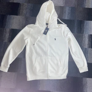 Vit hoodie från Polo Ralph Lauren - Snygg vit hoodie från Polo Ralph Lauren med dragkedja och klassisk logga broderad på bröstet. Tröjan har huva med snörning, känguruficka och ribbade muddar. Tillverkad i mjuk bomull, perfekt för en clean och stilren look.
