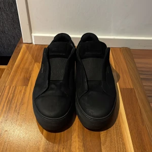 Svarta sneakers från Axel Arigato - Stilrena helsvarta sneakers från Axel Arigato med minimalistisk design och diskret logga i guld på sidan. Skorna är låga med rund tå och platt sula, tillverkade i skinn och har elastisk panel istället för snörning för en clean look.