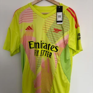 Arsenal målvaktströja Raya Adidas S - Arsenal målvaktströja från Adidas med Raya och nummer 22 på ryggen. Neon-gul bas med röda och rosa detaljer, röda ränder på axlarna och klubbens kanonmärke broderat på bröstet. Tillverkad i lätt och ventilerande polyester, perfekt för match eller träning.