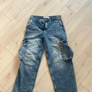 Blå cargojeans från Bershka - Säljer ett par blå cargojeans från Bershka med stora sidofickor och avslappnad passform. Jeansen har klassisk knappgylf, bälteshällor och är tillverkade i jeansmaterial. Perfekta för en chill och trendig look.