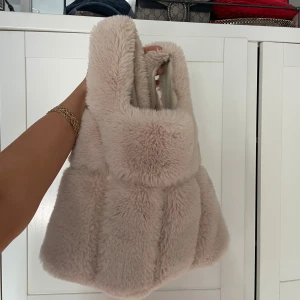 Beige fluffig axelväska i fuskpäls - Supermjuk och fluffig axelväska i beige fuskpäls med breda handtag. Väskan har en pösig form och stängs med magnetknapp. 
