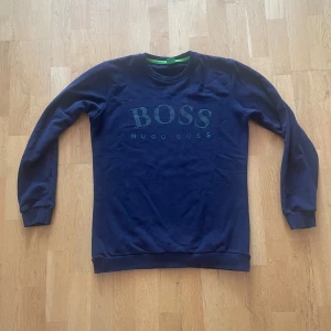 Mörkblå sweatshirt från Hugo Boss - Snygg mörkblå sweatshirt från Hugo Boss med stort grönt BOSS-tryck på bröstet. Tröjan har rund hals, ribbade muddar och är långärmad. Perfekt för dig som gillar stilrena och klassiska plagg med en modern touch. Passar en som är emellan 170-180 cm. Hör av er vid intressen! 