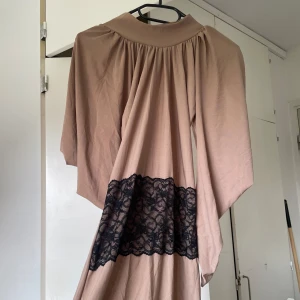 Beige långklänning med spetsdetalj - Långklänning i beige med bred svart spetsdetalj över midjan. Klänningen har långa ärmar och ett mjukt, fallande tyg som ger en avslappnad men elegant look. Perfekt för dig som gillar unika detaljer och klassisk look. 