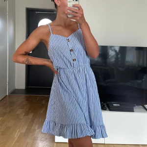 Blåvit randig sommarklänning - Söt blå och vit randig kortklänning med smala axelband, knappar framtill och volangkant nertill. Klänningen har en markerad midja och är perfekt för varma dagar. Lätt och luftig känsla med klassisk sommarstil.