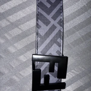 Svart Fendi bälte med FF-logga - Snyggt svart bälte från Fendi med ikoniskt FF-mönster längs hela bandet och en stor, matt svart spänne i metall. Bältet har en stilren och modern vibe, perfekt för att lyfta vilken outfit som helst. Materialet är skinn med präglat mönster.