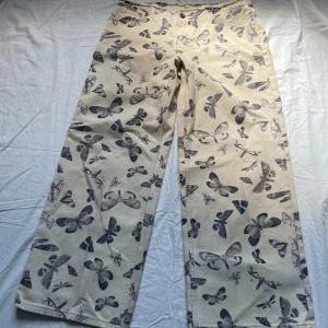 Wide baggy jeans med fjärilsmönster från Milk It - Snygga beige baggy jeans från Milk It med svart fjärilsmönster över hela byxan. Rå kant upptill för en edgy look. Tillverkade i bomull och har klassisk femficksdesign. Perfekta för dig som vill sticka ut med coola detaljer. Aldrig använda pga för korta för mig! 