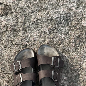 Bruna sandaler med dubbla spännen - Snygga bruna sandaler med två breda remmar och silvriga spännen. Fotbädden är formad för extra komfort och sulan har korkdetaljer. Perfekta för varma dagar och chill häng utomhus.