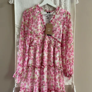 Blommig volangklänning från Vero Moda - Supersöt kort klänning från Vero Moda med rosa blommigt mönster på vit botten. Klänningen har långa ärmar, volangdetaljer och v-ringning med liten volangkant. Tillverkad i lätt polyester, perfekt för vår och sommar.