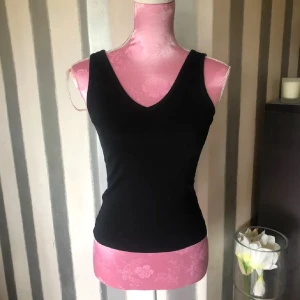 Svart ribbad linnetopp med v-ringning - En svart, ribbad linnetopp med v-ringning både fram och bak. Toppen är ärmlös och har en slim passform som sitter snyggt mot kroppen. Perfekt att styla med jeans eller kjol för en clean look.