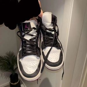 Nike Air Jordan 1 svart/vit/lila - Snygga Nike Air Jordan 1 sneakers i svart och vitt med lila detaljer. Klassisk high-top modell med svart snörning och ikonisk Air Jordan-logga på sidan. Ovandel i skinn och mocka. Använda men är fortfarande i fint skick, fick dem i present så vet ej vad dem köptes för men säljer dem för 900kr. 