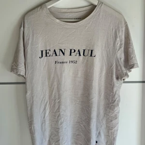 Beige t-shirt från Jean Paul - Snygg beige t-shirt från Jean Paul med svart tryck framtill där det står 'JEAN PAUL France 1952'. Klassisk rund halsringning och korta ärmar. Tillverkad i mjuk bomull som känns skön mot huden. Perfekt för en clean och stilren look.