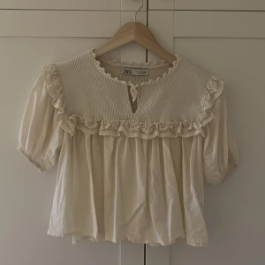 Beige volangblus från Zara - Supersöt beige blus från Zara med stickad överdel och volangdetaljer längs axlar och bröst. Puffiga korta ärmar och knytning vid halsen ger en romantisk vibe. Perfekt för dig som gillar vintageinspirerad stil och mjuka material.