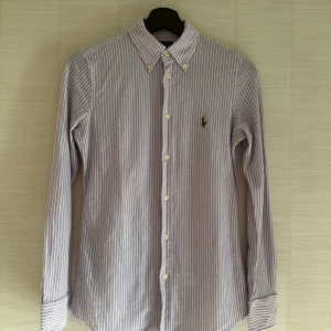 Randig skjorta från Ralph Lauren - Klassisk knit Oxford-skjorta från Ralph Lauren i ljuslila med tunna ränder. Elegant och tidlös. Storlek S men passar XS och XXS bättre 
