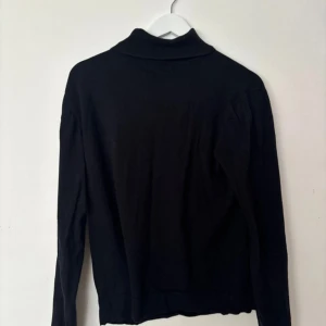Svart polotröja i bomull - En klassisk svart polotröja med lång ärm och ribbad krage. Tröjan är i mjuk bomull och har en enkel, clean look som funkar till allt. Perfekt för lager-på-lager och snygg till både jeans och kjol.