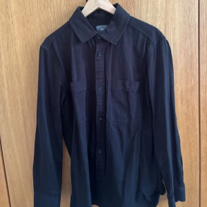 Svart overshirt från H&M i bomull - Svart overshirt från H&M i mjuk bomull. Skjortan har klassisk krage, två bröstfickor och knäppning framtill. Långärmad och med en rak, lite loose passform som funkar till många olika stilar.