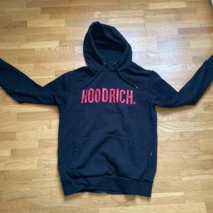 Svart HOODRICH hoodie med röd logga - Svart hoodie från HOODRICH i storlek small med stor röd logga framtill. Klassisk känguruficka, huva med snörning och långa ärmar. Mjuk insida och relaxed fit, perfekt för en streetwear-look.