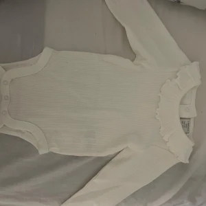 Vit ribbad body med volang från Lindex - Supersöt vit ribbad body från Lindex i storlek 62. Långärmad modell med volangdetalj runt halsen och praktiska tryckknappar i grenen. Perfekt för dig som gillar stilrena och gulliga babykläder,oanvänd