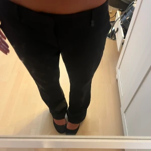 Svarta kostymbyxor med rak passform - Säljer ett par svarta kostymbyxor med klassisk rak passform och hällor för bälte. Byxorna har midwaist och är perfekta för en clean och stilren look. Materialet känns mjukt och följsamt, och de har diskreta fickor i sidorna. Säljer de då jag köpte för stor storlek!