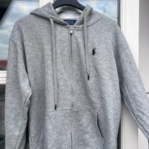 Grå hoodie från Polo Ralph Lauren - Snygg grå hoodie från Polo Ralph Lauren med dragkedja och huva. Klassisk svart broderad logga på bröstet, stora fickor framtill och justerbara snören i huvan. Mjuk bomullsblandning som är perfekt för chill dagar.