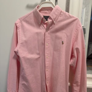 Rosa skjorta från Polo Ralph Lauren - Klassisk rosa skjorta från Polo Ralph Lauren med button-down krage och broderad logga på bröstet. Skjortan har långa ärmar och knäppning framtill. Perfekt för en stilren och fräsch look.