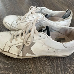 Golden Goose vita sneakers med stjärna - Golden Goose sneakers i vitt skinn med ikonisk grå stjärna på sidan och GGDB-logga på plösen. Skorna har snörning, rund tå och slitna detaljer för en cool, vintage look. Perfekta för dig som gillar streetstyle och unika detaljer. Rejält använda 