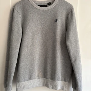 Grå sweatshirt från G-Star RAW - En stilren grå sweatshirt från G-Star RAW med liten logga på bröstet. Tröjan har rund halsringning, ribbade muddar och är tillverkad i mjuk bomullsmix. En svag missfärgning i nederkant på tröjan, ses site bilden. Annars jättefint skick. 