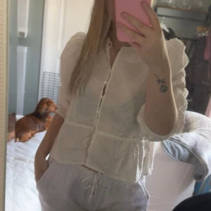 Ljusvit blus med volang och knappar - Söt och luftig ljusvit blus med volangdetaljer och knappar framtill. Blusen har långärmade ärmar med lätt puff och är tillverkad i ett tunt, transparent material som ger en romantisk känsla. Perfekt för dig som gillar en feminin och trendig stil.