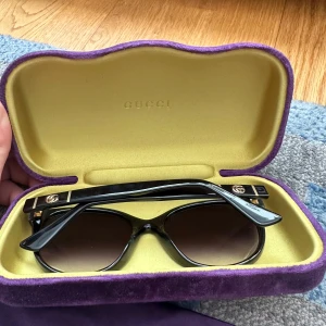 Svarta cat eye solglasögon från Gucci - Säljer ett par svarta cat eye solglasögon från Gucci med snygga gulddetaljer på skalmarna och GG-logga. Kommer med originalfodral i lila och gul sammet, putsduk och tygpåse. Perfekta för att lyfta din stil till nästa nivå.