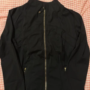 Lululemon define jacket gold zipper - Svart träningsjacka från Lululemon med snygga guldiga dragkedjor och diskreta sömdetaljer. Jackan har hög krage, långa ärmar och praktiska fickor med dragkedja. Perfekt för dig som gillar sportig och clean stil.