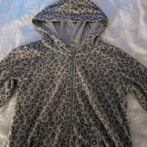 Grå leopardmönstrad hoodie med dragkedja - Grå hoodie med leopardmönster, hel dragkedja framtill och huva. Tröjan har långa ärmar och känguruficka, perfekt för en chill och trendig look. Mjuk bomullskänsla och relaxed passform.