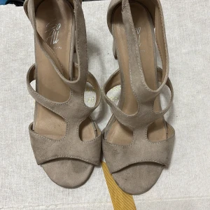 Beige mockasandaler med klack - Snygga sandaler i beige mocka med öppen tå och blockklack. Skorna har coola utskurna detaljer och dragkedja bak för enkel påtagning. Perfekta för dig som gillar stilrena och trendiga skor med en twist.