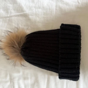 Svart ribbad mössa med pälsboll - Svart stickad mössa med bred uppvikt kant och en stor, fluffig pälsboll i beige nyans på toppen. Klassisk ribbstickning ger en snygg och enkel look, perfekt för kalla dagar. Pälsbollen går även att ta av!