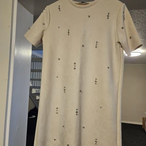 Beige kortklänning med pärldetaljer - Stilren beige kortklänning med korta ärmar och rund hals. Klänningen är dekorerad med små strassliknande stenar som ger en glittrig touch. Materialet känns mjukt och har en lätt struktur. Perfekt för dig som vill ha något enkelt men ändå extra. Oanvänd fast utan prislapp då det var en present. 