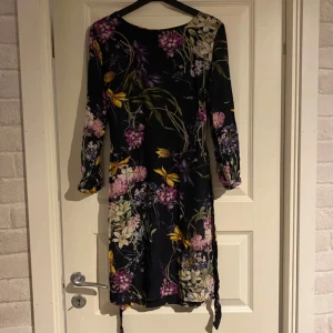Klänning - Säljer en vacker blommig klänning från Hansen o Jacob i storlek XS. Klänningen har ett mörkt blommönster med inslag av lila, rosa och gult. Den är medellång i ärmarna och har en elegant passform som passar perfekt för speciella tillfällen. Klänningen är tillverkad i ett mjukt och bekvämt material. Den är endast testad, se sista bilden för defekt, en av knapparna har orsakat lite uppslitningar. Låtsas knappar på baksidan och äkta på ärmarna. Nypris 2800kr
