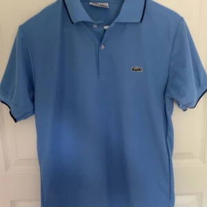 Blå pikétröja från Lacoste - Snygg ljusblå pikétröja från Lacoste med klassisk krage och två knappar framtill. Tröjan har korta ärmar med mörkblå kant och den ikoniska krokodilloggan på bröstet. Mycket bra skick! Skriv vid minsta lilla fundering!