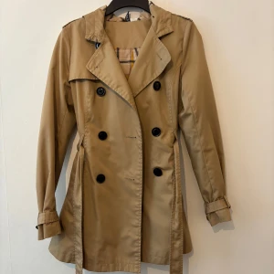 Beige trenchcoat med huva och bälte - Stilren beige trenchcoat från C&A med dubbelknäppning, bälte i midjan och justerbara ärmslut. Jackan har en avtagbar huva och rutigt innerfoder som ger en snygg detalj. Perfekt för dig som vill ha en klassisk look med modern twist.