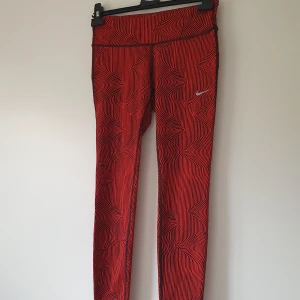 Nike Dri-Fit leggings röd/svart S - Nike Dri-Fit leggings i rött med svarta vågiga linjer. Tight passform, hög midja och liten dragkedjeficka bak. Perfekta för träning med sitt stretchiga och fuktavvisande material. Snygg och sportig look med Nike-logga på låret.  Lapparna bort klippta, små noppror