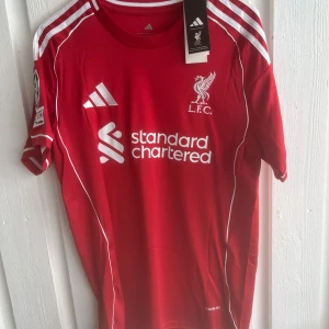 Liverpool FC röd matchtröja Adidas - Liverpool FCs officiella röda matchtröja från Adidas med vita detaljer och tryck. Kortärmad modell med klubbmärke, sponsorlogga och Champions League-märke på ärmen. Namn och nummer Frimpong 30 på ryggen. Tillverkad i lätt funktionsmaterial.